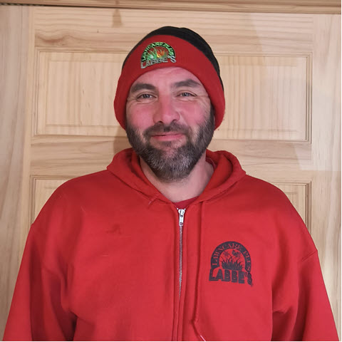 A bearded man in a red hoodie and beanie stands in front of a wooden door, perhaps enjoying some rare free time away from his duties at Labbe's Lawn Care Plus.