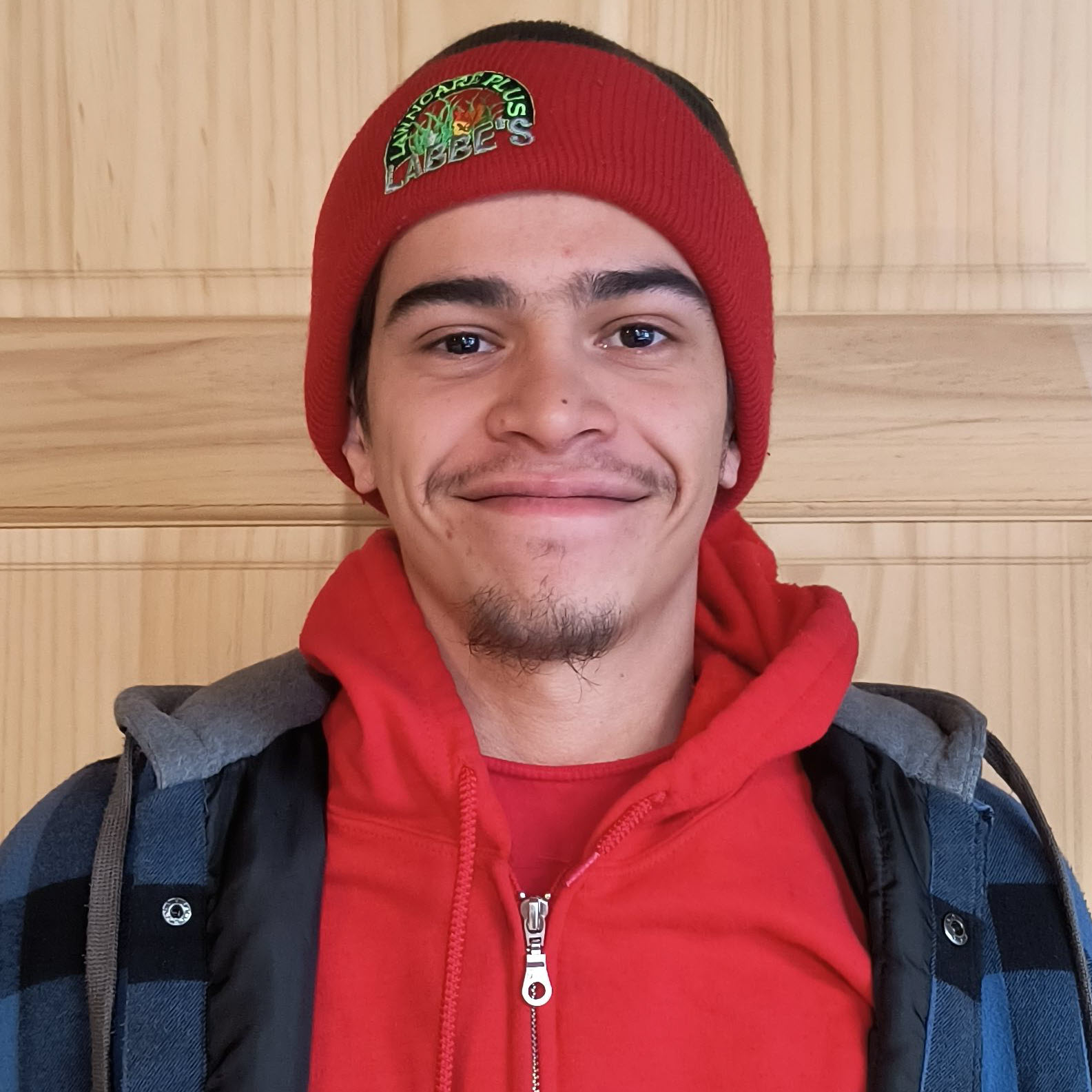 A person wearing a red beanie and hoodie smiles during their free time, standing against a wooden background.