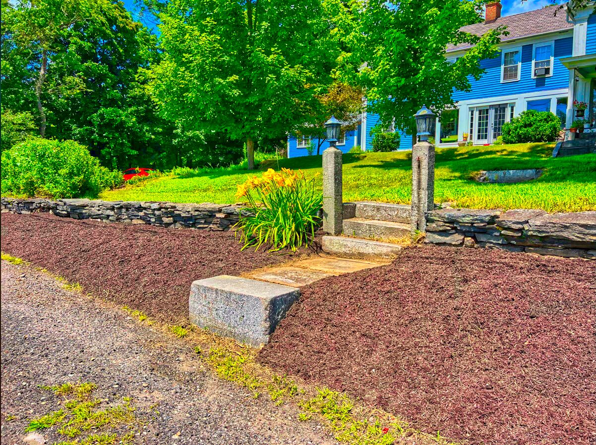 Stone steps lead through a mulched garden to a blue house with a porch. Trees and greenery surround the area. Stone steps lead through a mulched garden to a blue house with a porch. Trees and greenery surround the area.